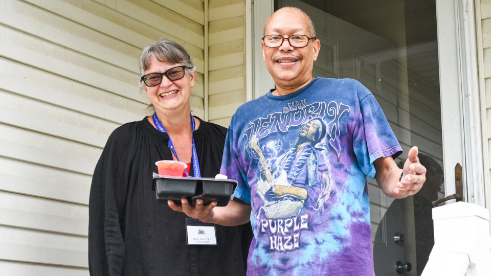 Male client on right smiling with hands out and meal tray in right hand next to his volunteer on his right smiling in front the client's front doorn