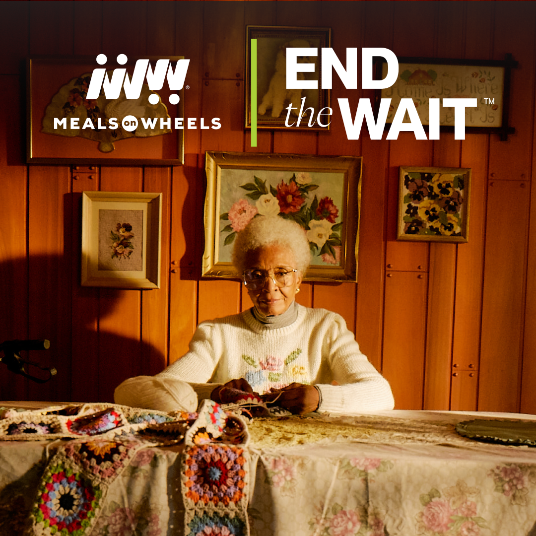 Meals on Wheels client sitting at her dinner table with the text, End the Wait at the top