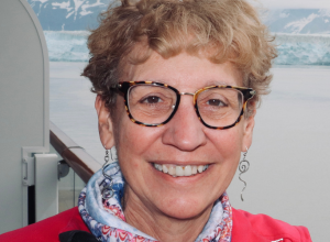 Headshot of Jeanne Ryan smiling with a pair of glasses on, and multicolored scarf, red top, and dangling earrings.