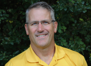 Kevin Dougherty smiling with glasses on standing in front of small tree leaves wearing a yellow CertaPro Painters polo shirt.